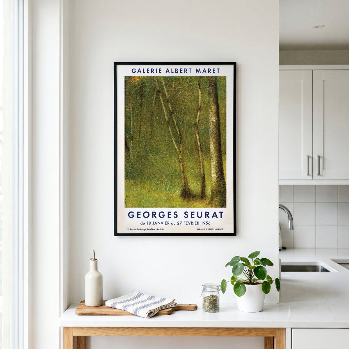 A framed Georges Seurat forest art print hangs on a white kitchen wall above a countertop with a bottle, towel, and small plant.