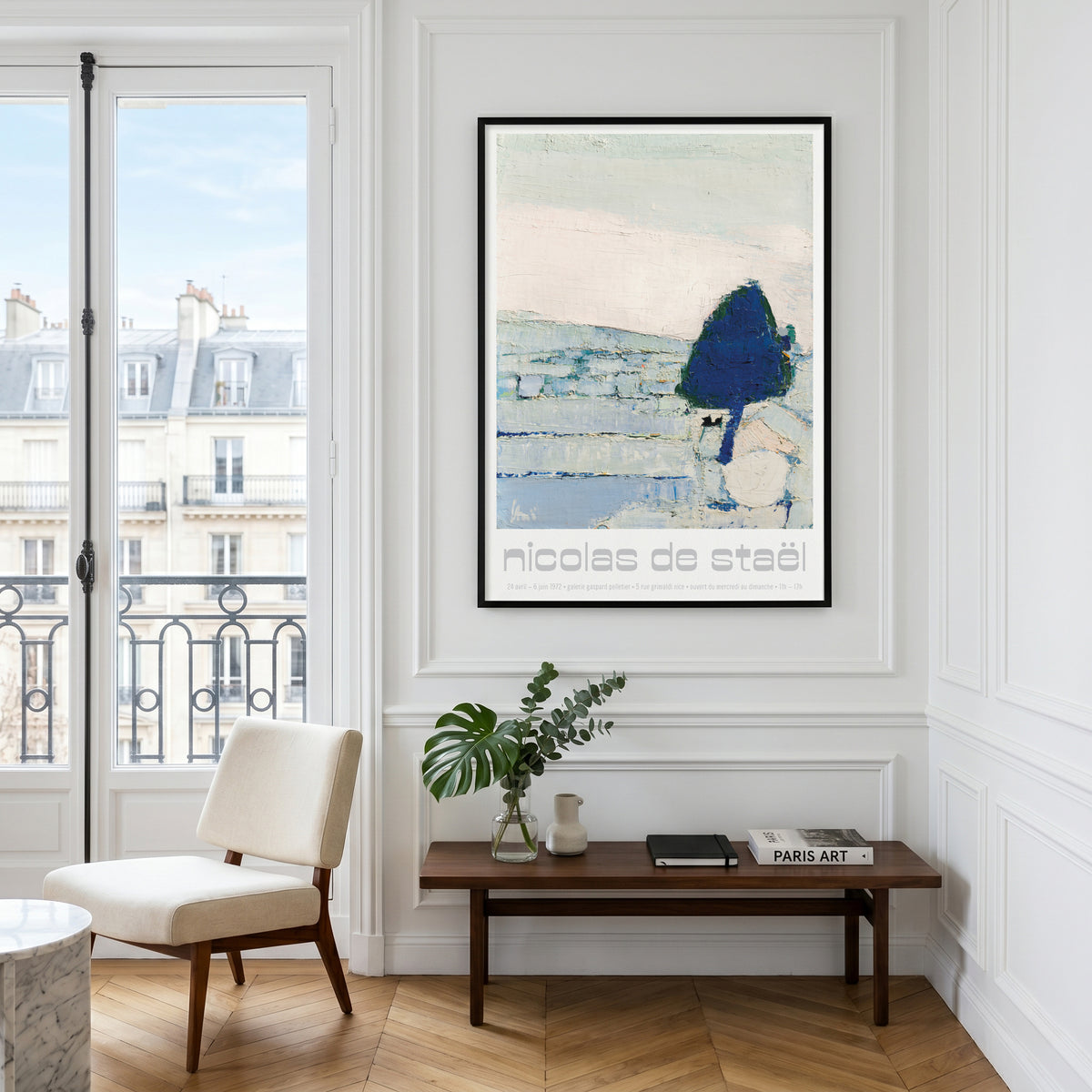 A framed art print featuring an abstract landscape with a dark blue tree and the text 'Nicolas de steel' is displayed on a white paneled wall in a bright room with a window, chair, and bench.