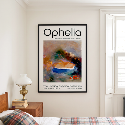A framed art print titled 'Ophelia' hangs on a cream-colored wall in a bedroom setting, next to a window and a wooden bedside table with a lamp.
