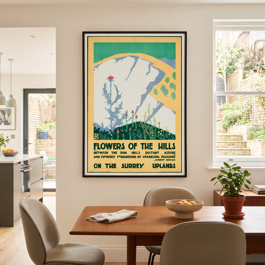 A framed vintage-style travel poster hanging on a beige wall above a wooden dining table in a modern kitchen and dining room setting. The poster reads 'Flowers of the Hills' and features a stylized illustration of a white cliff with green hills and flowers.