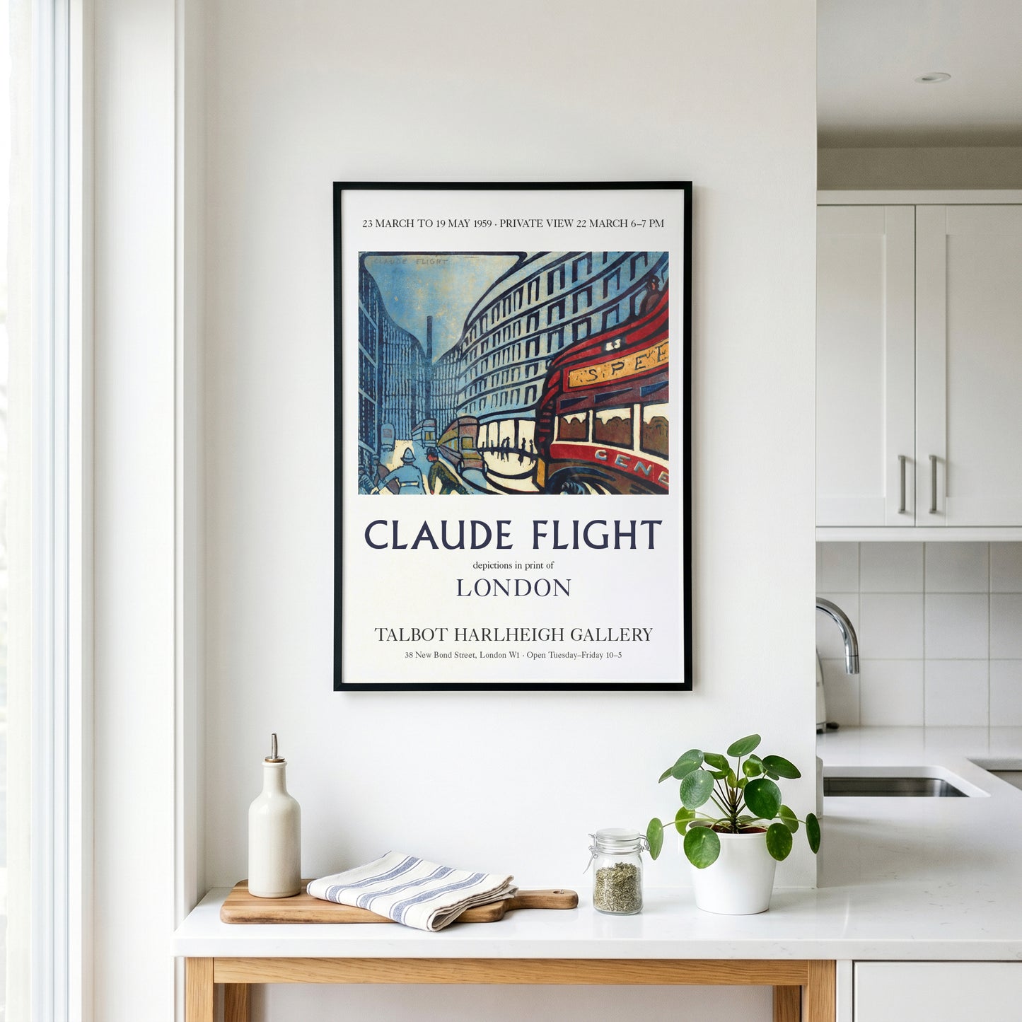 A framed vintage-style travel poster for Claude Flight in London hangs on a white kitchen wall above a countertop with a bottle, cloth, and small plant.
