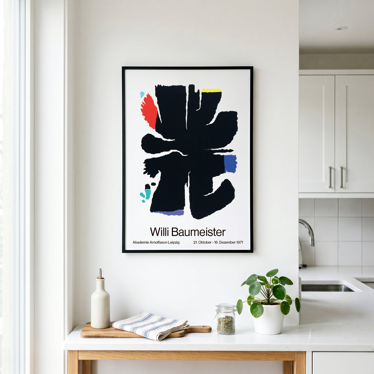 A framed art print featuring an abstract black brushstroke design with the text 'Willi Baumeister' and dates, displayed on a white kitchen wall above a countertop with a plant and small bottles.