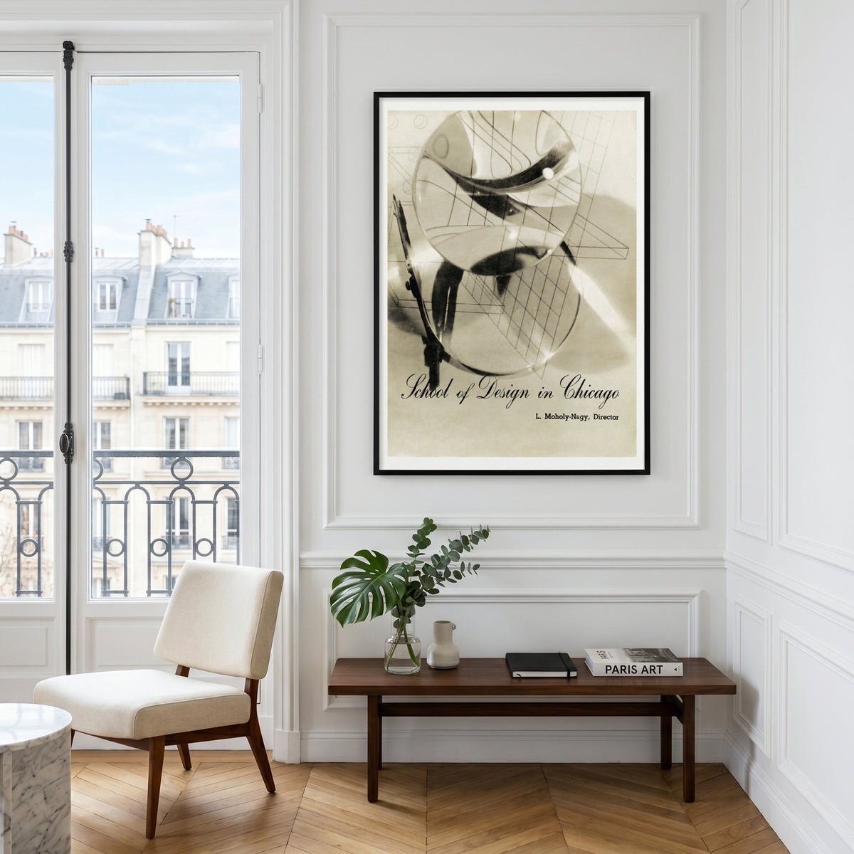 A framed art print featuring an abstract design with the text 'Design of Living in Chicago' and 'L. Mies van der Rohe' is displayed on a white wall in a bright, modern room with a wooden bench, chair, and window view.