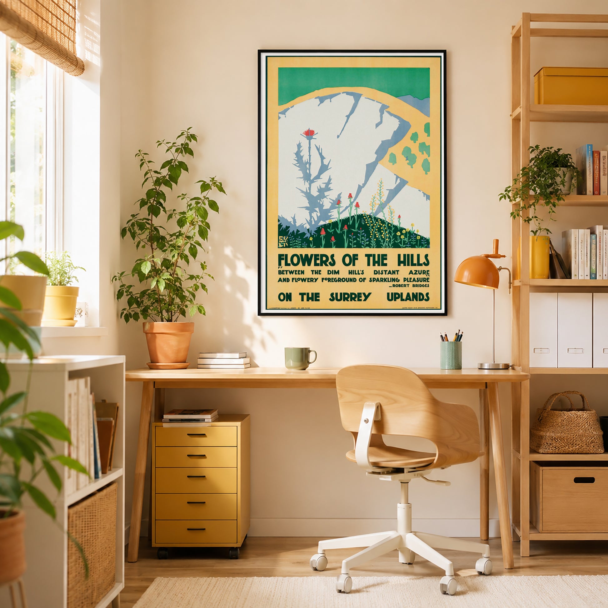 A framed vintage-style travel poster hangs on a beige wall above a wooden desk in a bright home office. The poster reads 'Flowers of the Hills' and features a teal, yellow, and green landscape illustration.