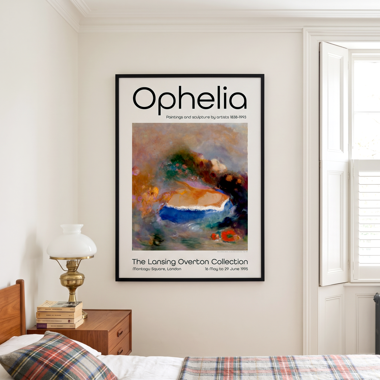 A framed art print titled 'Ophelia' hangs on a cream-colored wall in a bedroom setting, next to a window and a wooden bedside table with a lamp.