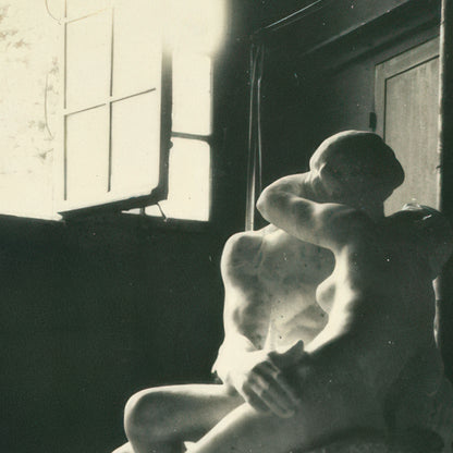 A black and white photograph of a seated nude female figure in a dim interior, with strong sunlight streaming through a window and a door in the background.