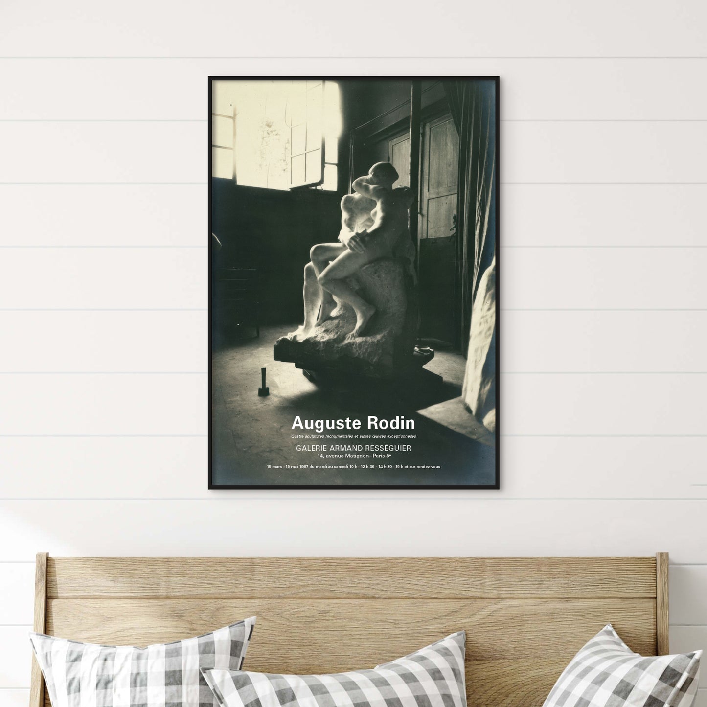 A framed black and white art print of Auguste Rodin's sculpture 'Galérien Armado' displayed on a white shiplap wall above a wooden headboard with gingham pillows.