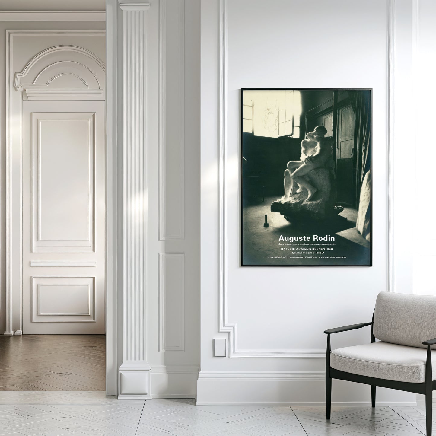 A framed black and white poster of Auguste Rodin hanging on a white wall in a classic European hallway, next to a light gray armchair.