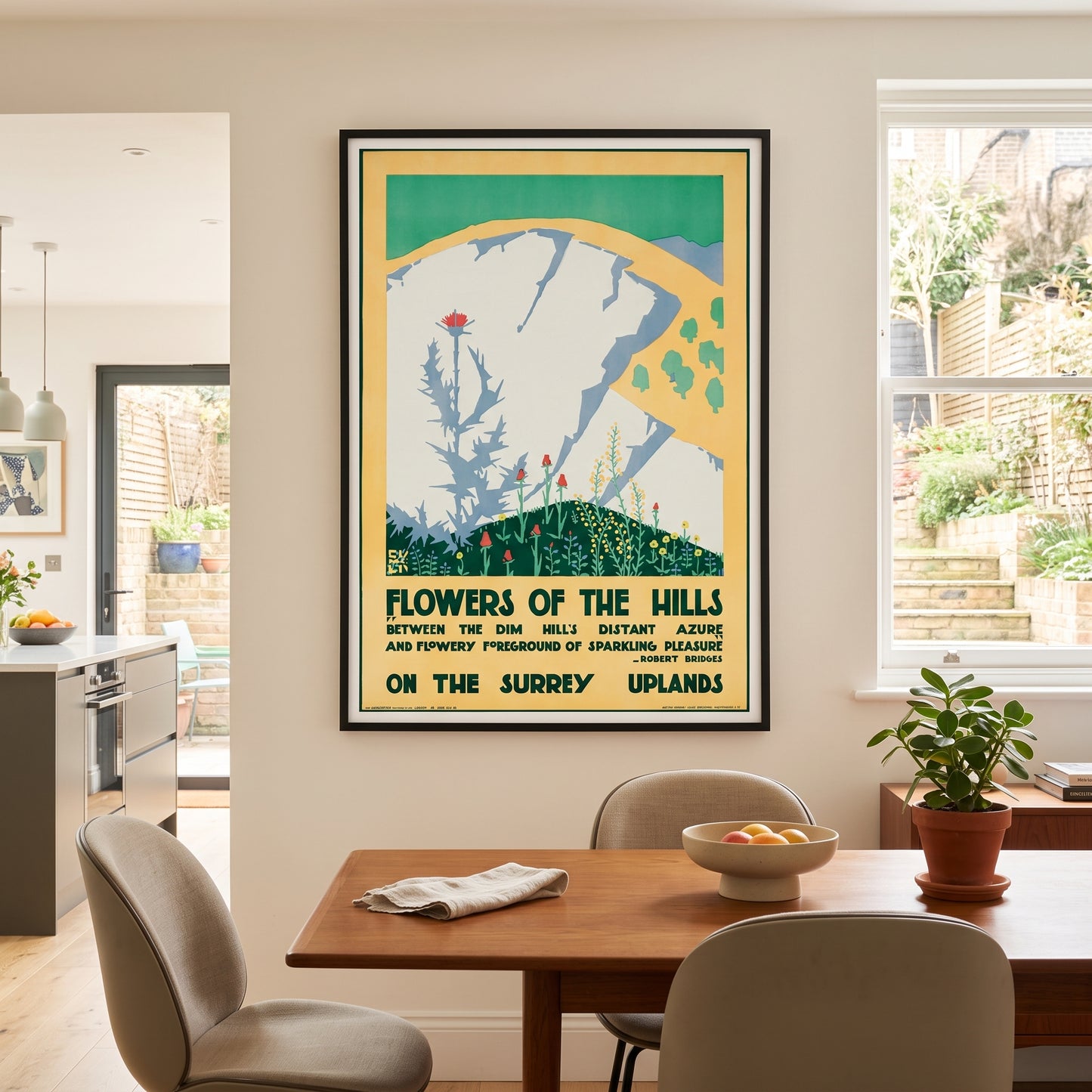 A framed vintage-style travel poster hanging on a beige wall above a wooden dining table in a modern kitchen and dining room setting. The poster reads 'Flowers of the Hills' and features a stylized illustration of a white cliff with green hills and flowers.