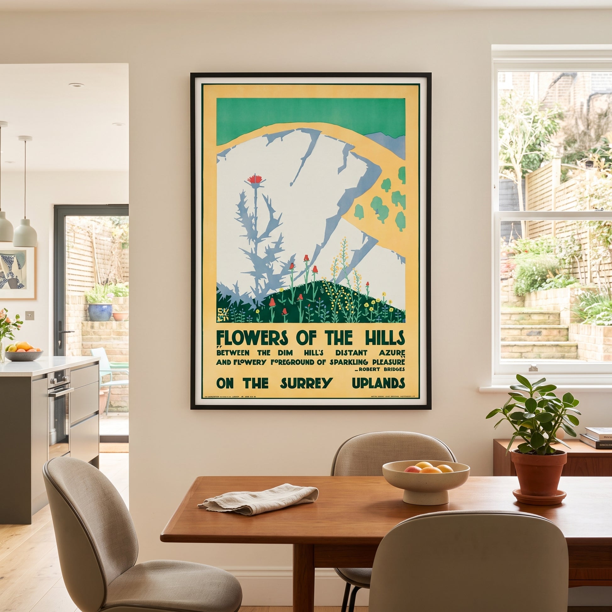 A framed vintage-style travel poster hanging on a beige wall above a wooden dining table in a modern kitchen and dining room setting. The poster reads 'Flowers of the Hills' and features a stylized illustration of a white cliff with green hills and flowers.
