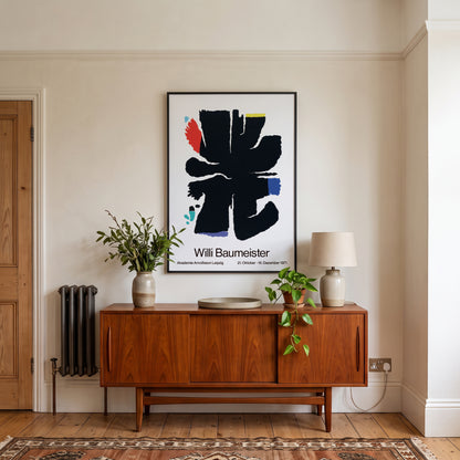 A framed art print of Willi Baumeister's abstract black brushstroke design hangs on a cream-colored wall above a mid-century modern wooden credenza in a styled room setting.