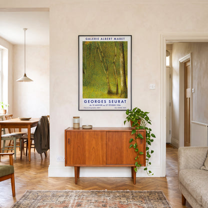 A framed vintage-style art print of a green forest by Georges Seurat hangs on a beige wall in a modern living room setting above a mid-century wooden sideboard with a trailing green plant.